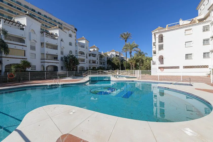Una piscina se encuentra entre edificios de apartamentos blancos bajo un cielo azul despejado.