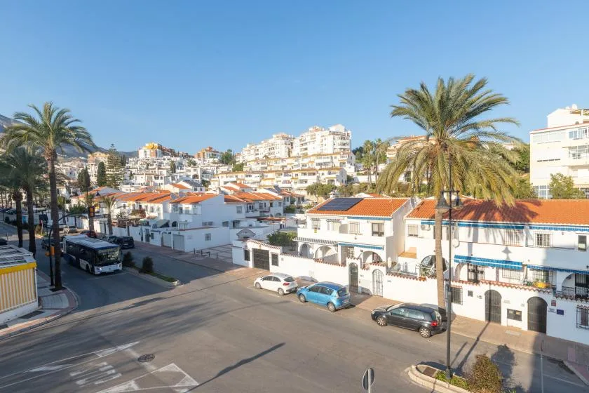 A sunny street lined with white buildings, parked cars, and palm trees.