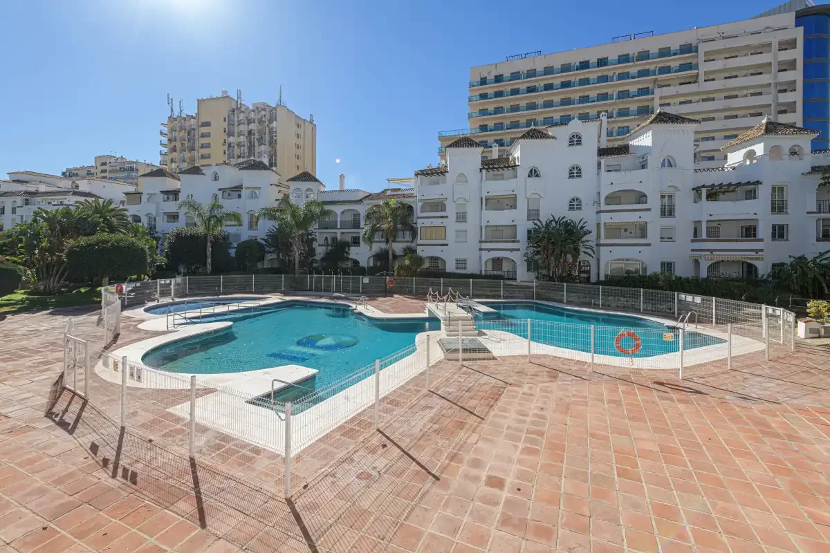 An outdoor swimming pool sits beside white apartment buildings under a clear sky.