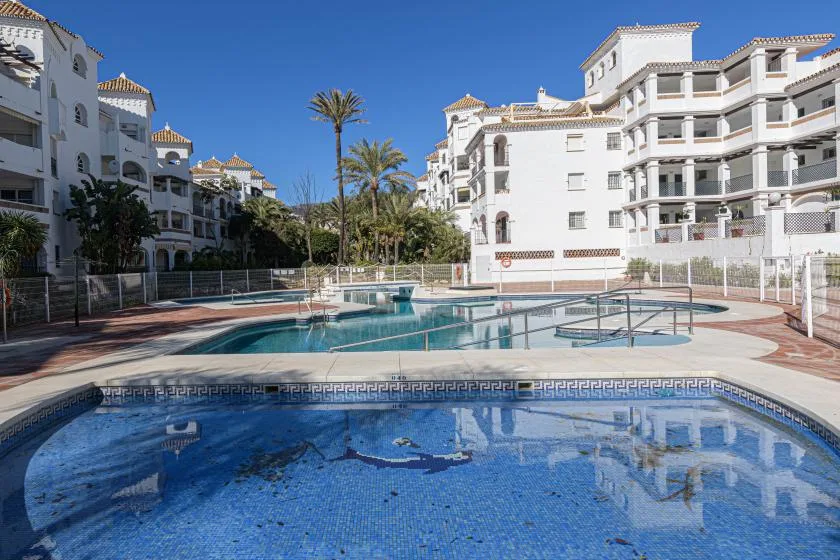 An outdoor swimming pool sits among white apartment buildings and palm trees.