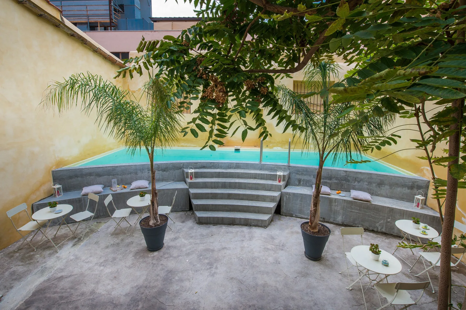 A courtyard pool with tiered steps, potted palms, and small seating areas.