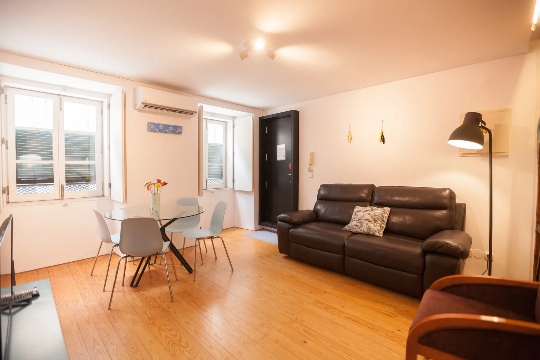A bright living and dining area with a dark sofa, glass table, and windows.