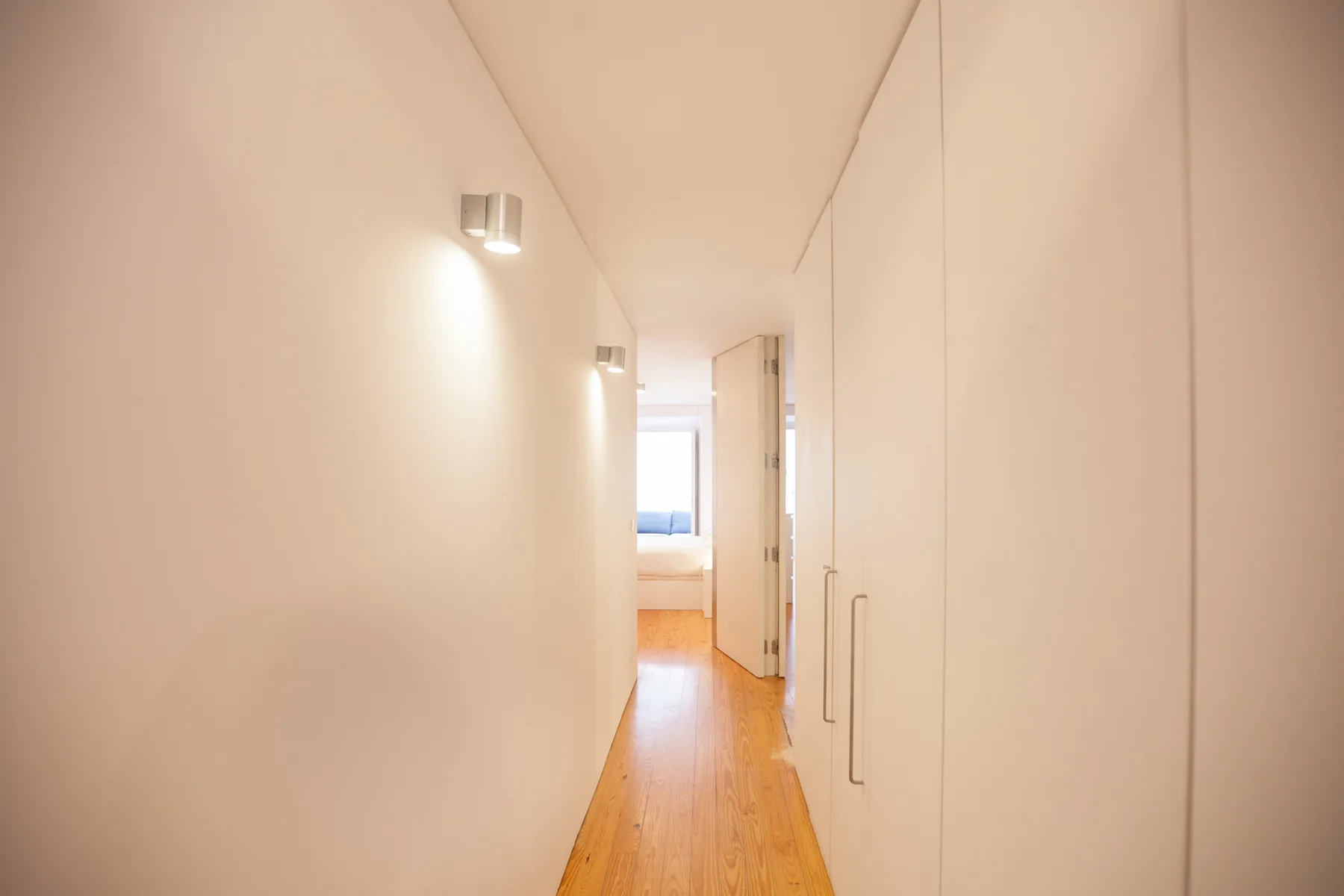 A narrow hallway with wood flooring leads toward a bright room.