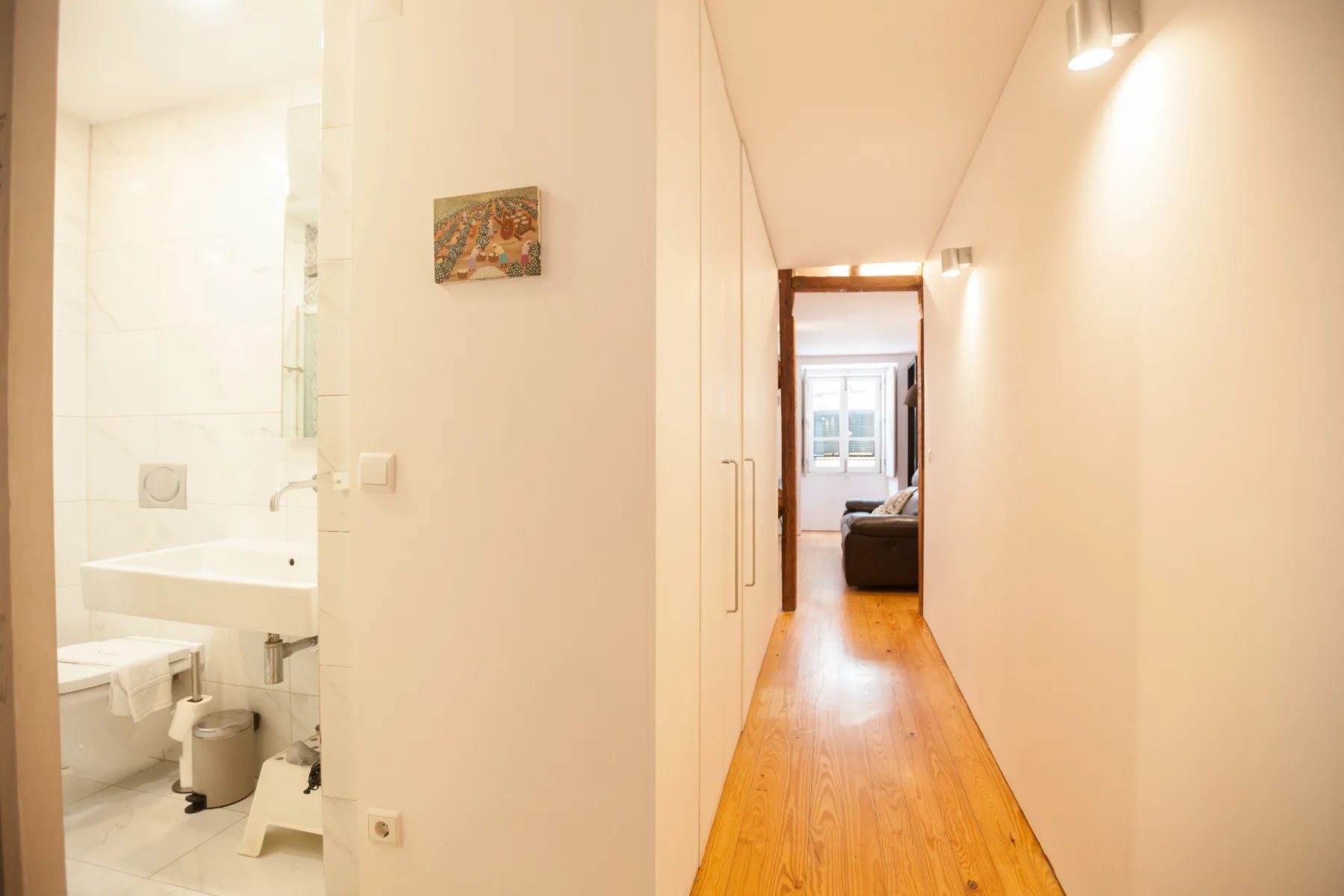 A bright apartment hallway with wooden flooring leads toward a bedroom.