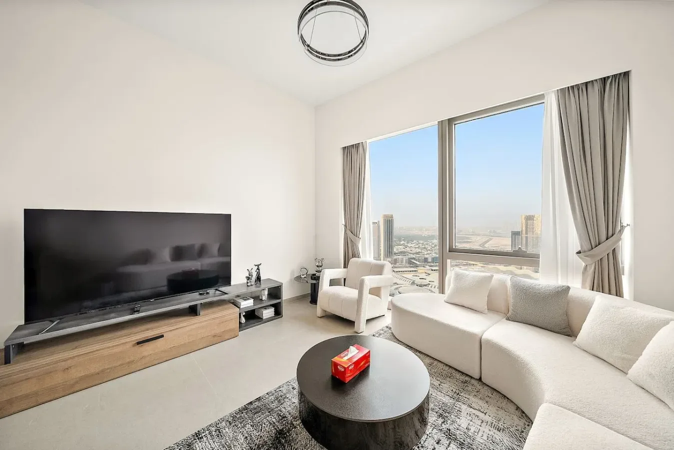 Una sala de estar luminosa con asientos, un televisor y grandes ventanas con vistas a la ciudad.