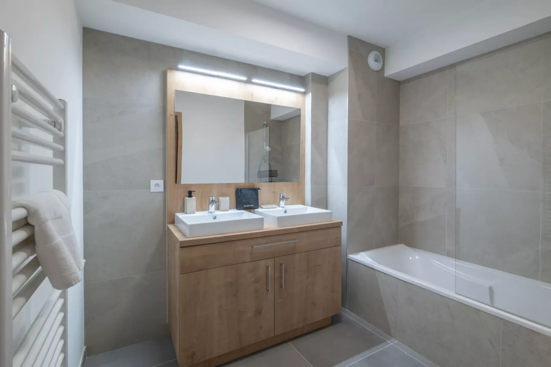 A tiled bathroom with a wooden vanity, double sinks, and a bathtub.