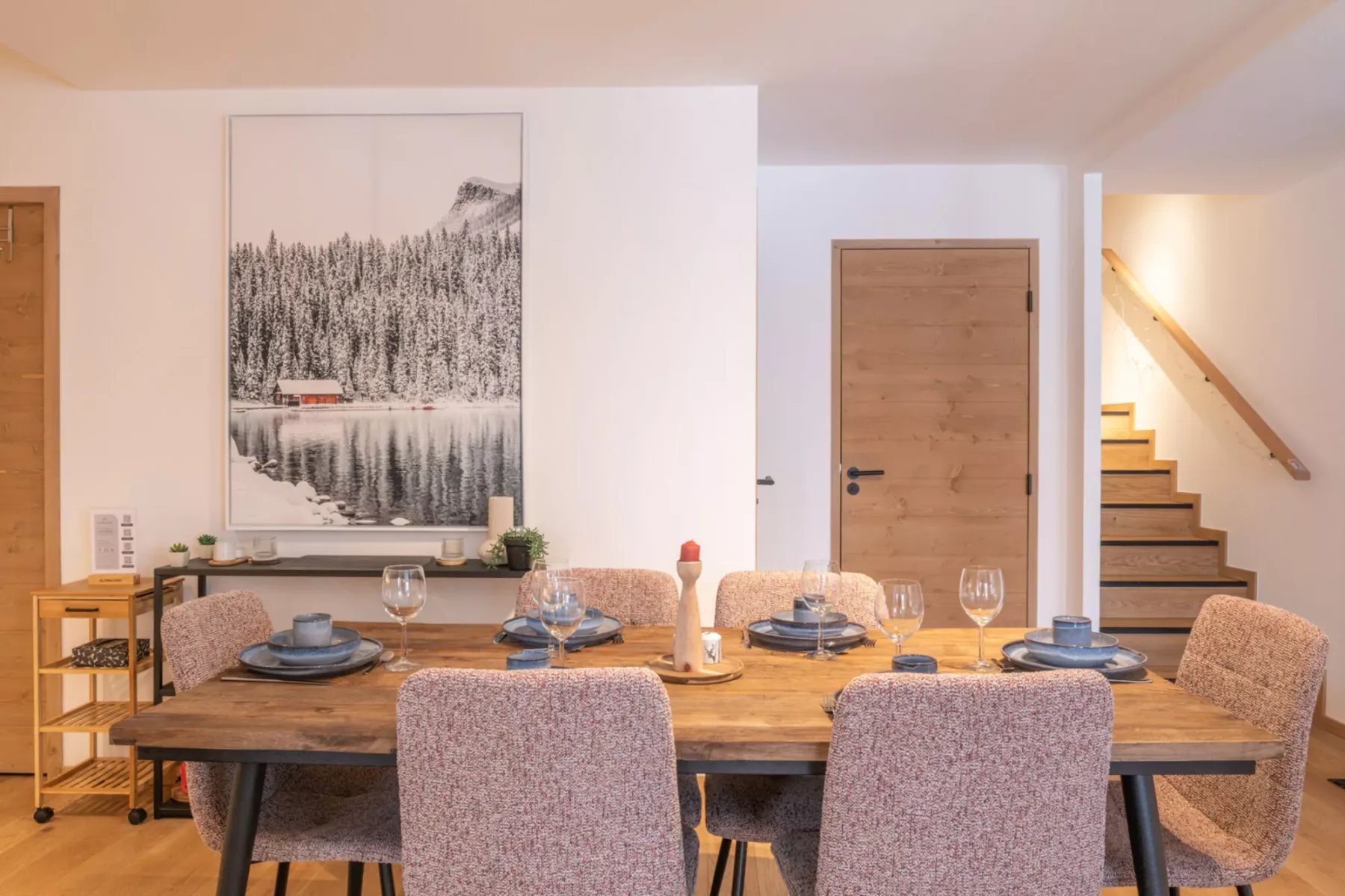 A dining area with a wooden table set with dishes and a large landscape print on the wall.