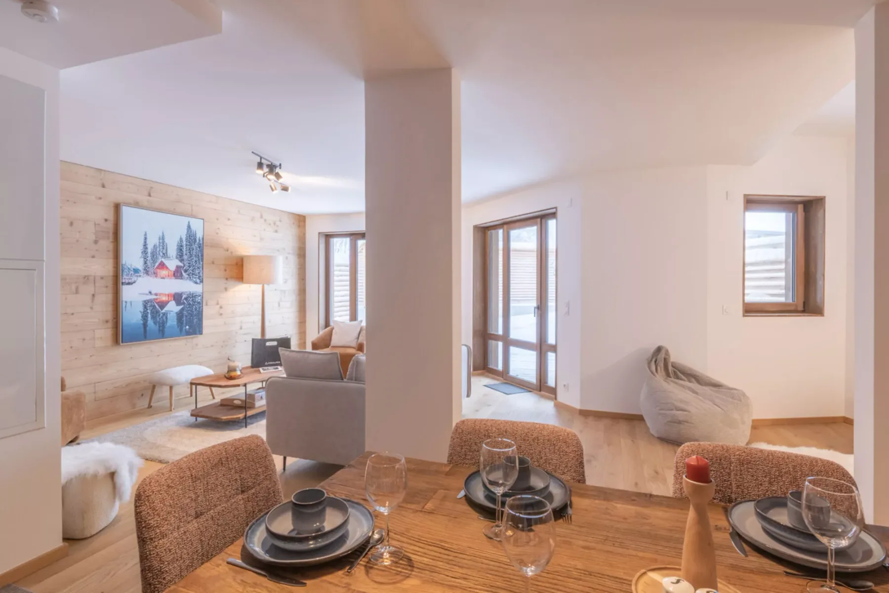 A dining area opens onto a living room with large windows and light wood walls.