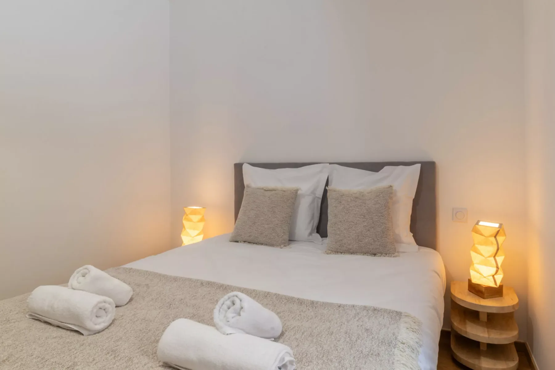 A bedroom with a neatly made bed, folded towels, and warm bedside lighting.