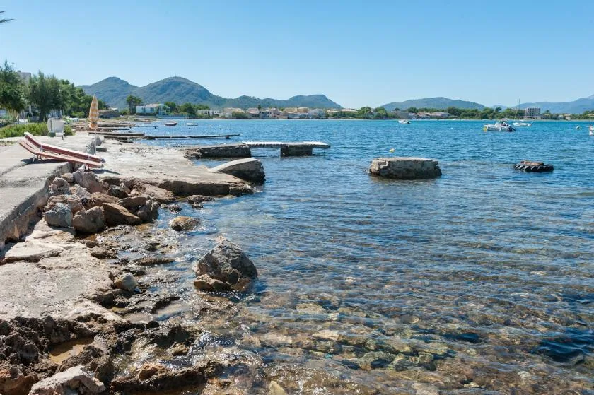 A rocky shoreline meets clear blue water, with distant hills across the bay.