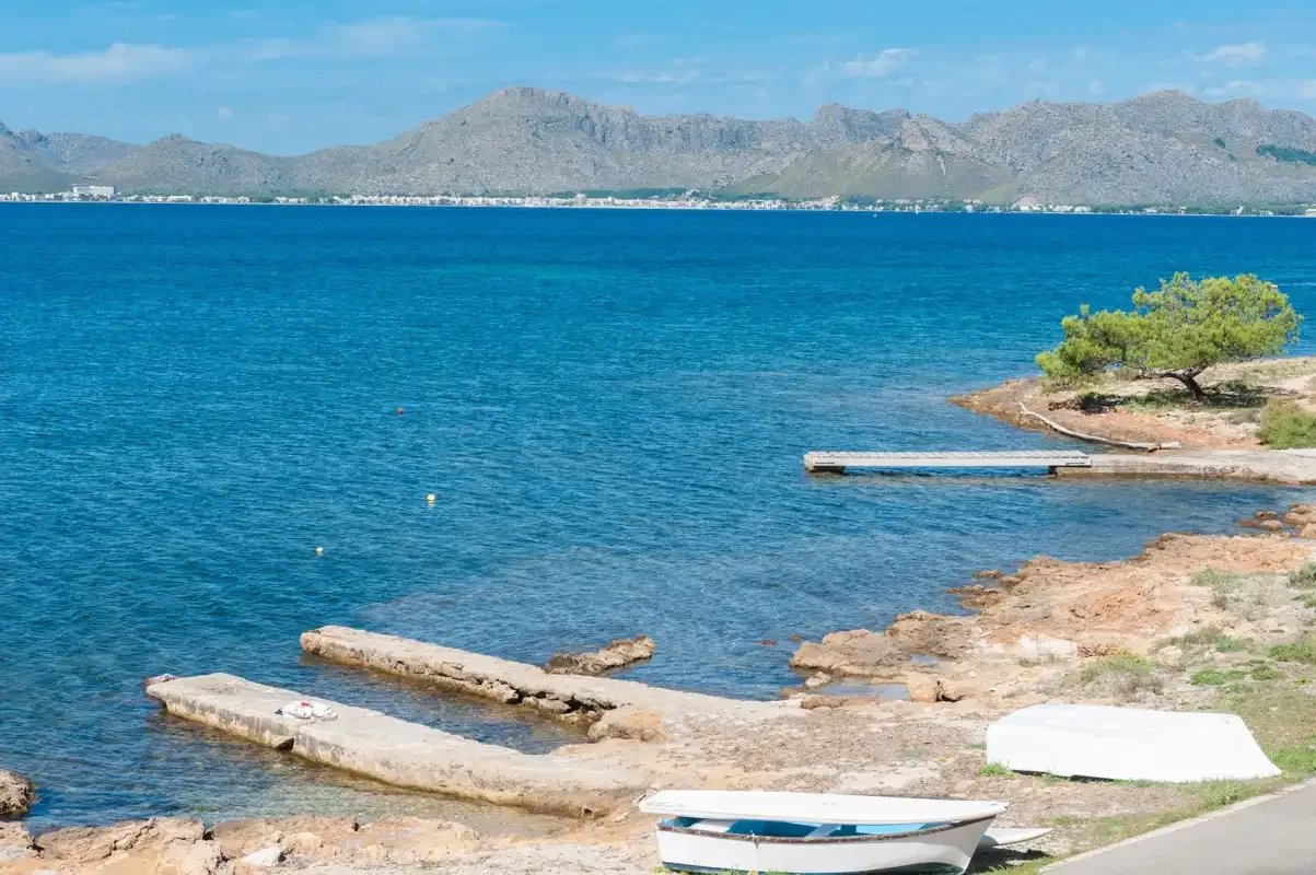 A rocky shoreline borders clear blue water, with mountains visible across the bay.