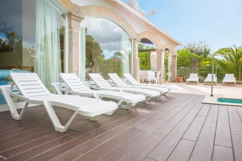 A poolside patio has white loungers beside arched columns and glass doors.