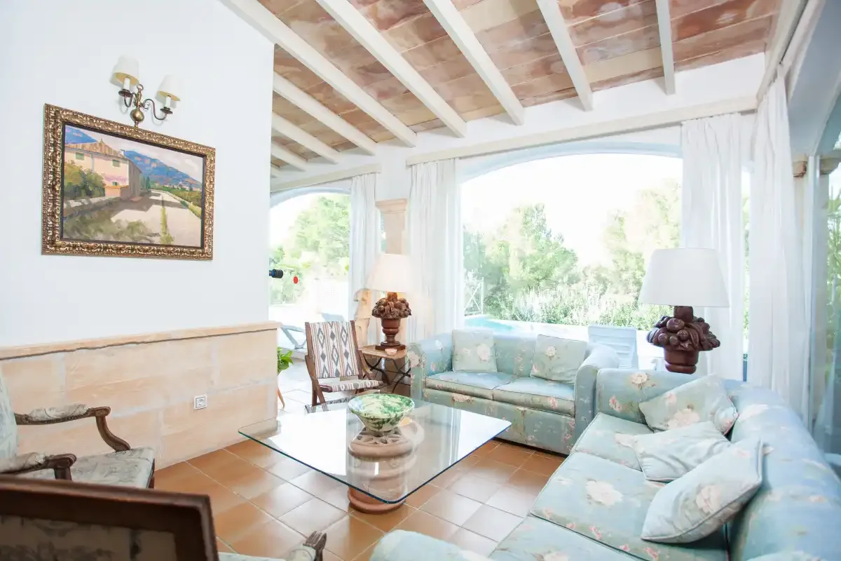 A bright living room with pale seating, a glass coffee table, and large windows.