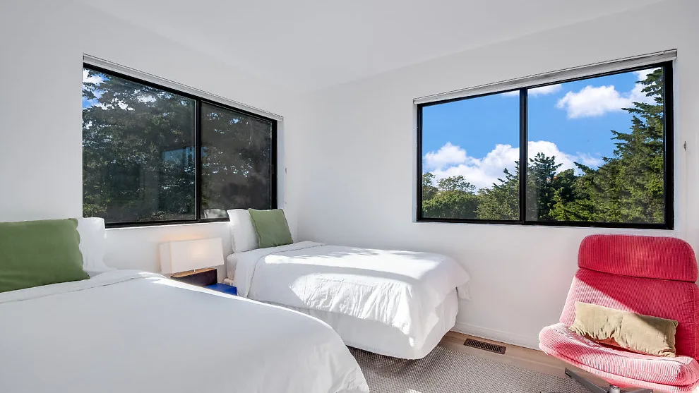Un dormitorio luminoso con dos camas y grandes ventanas con vista a los árboles.