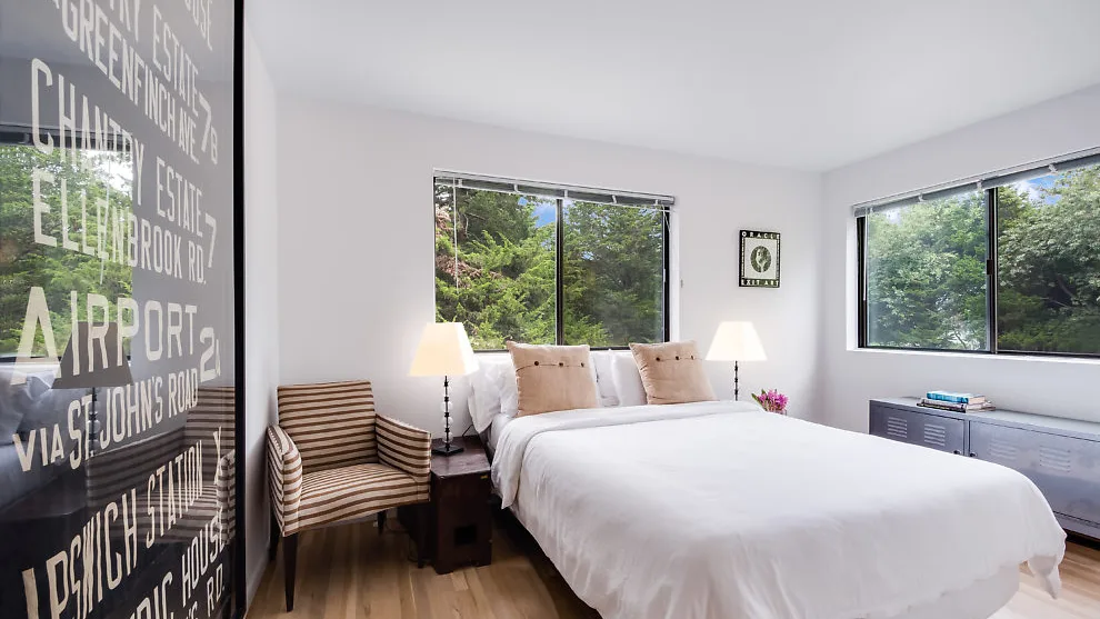 Un dormitorio luminoso con una cama bien tendida y grandes ventanas con vistas a la vegetación.