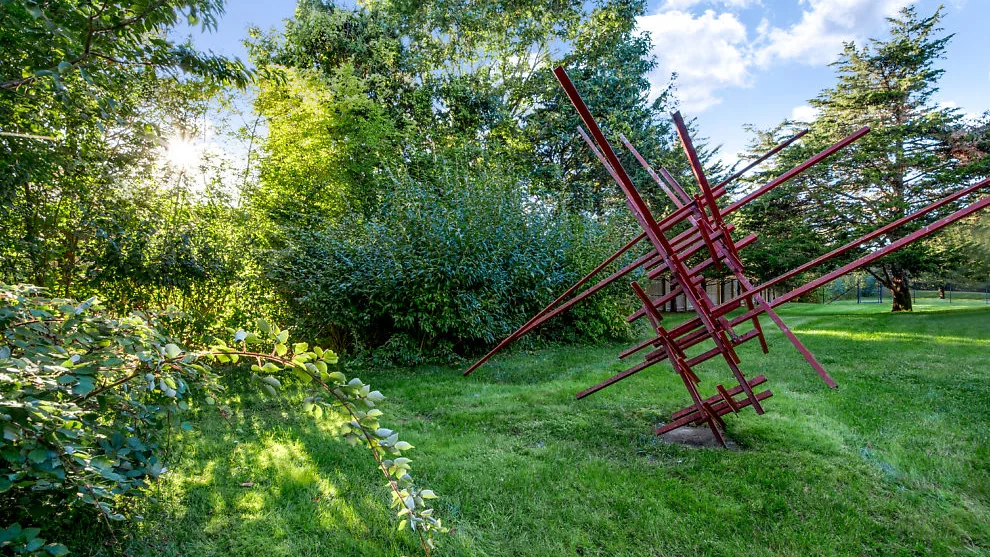 Una escultura de metal roja se alza sobre un césped entre árboles.