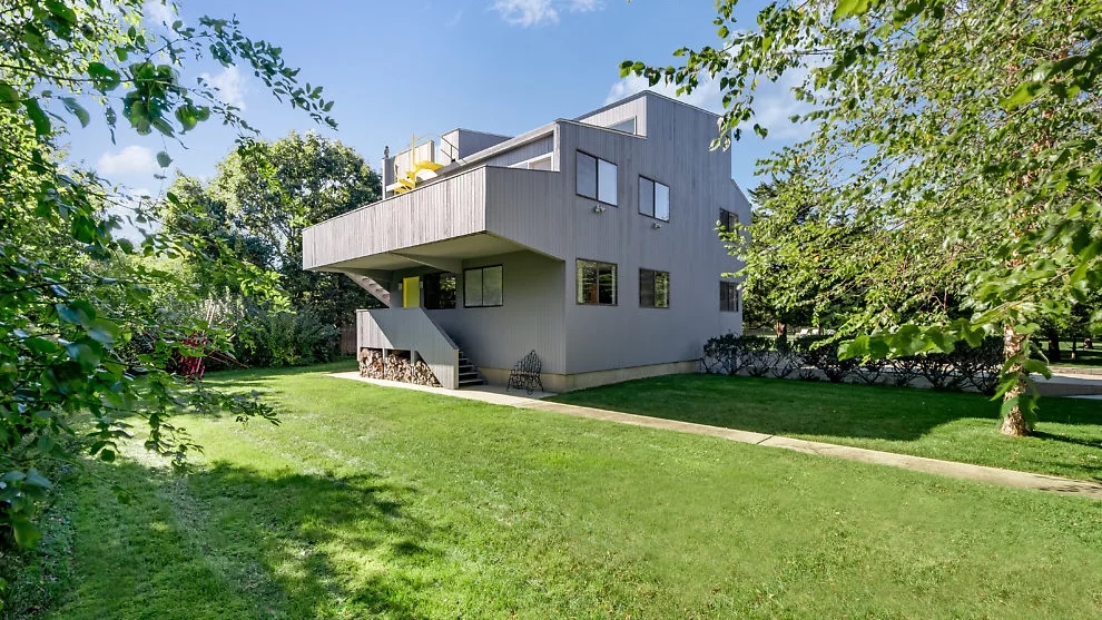 Una casa moderna se encuentra junto a un amplio césped, rodeada de árboles y vegetación.