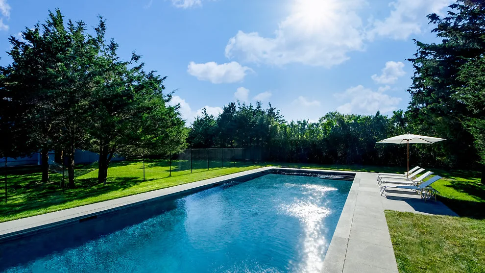 An outdoor swimming pool sits beside a grassy lawn and trees.