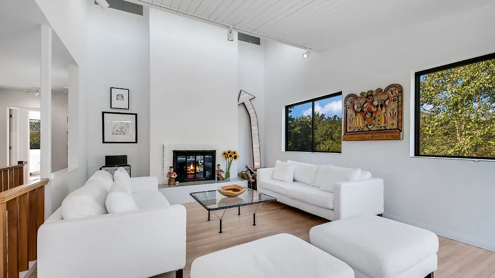 Una sala de estar luminosa con asientos blancos, una mesa de centro de vidrio y ventanas grandes.