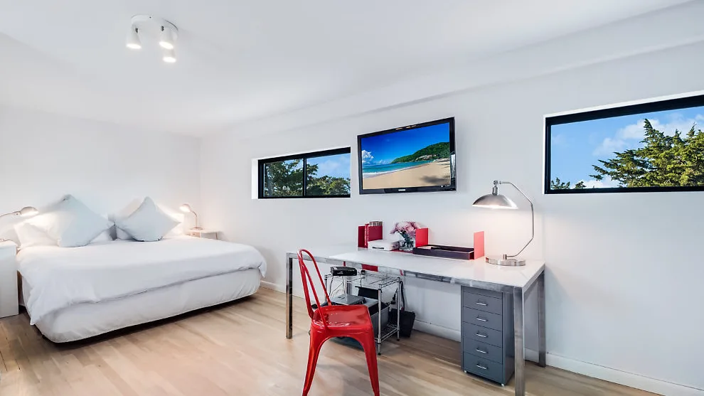 Un dormitorio luminoso con una cama bien tendida y un escritorio bajo las ventanas.