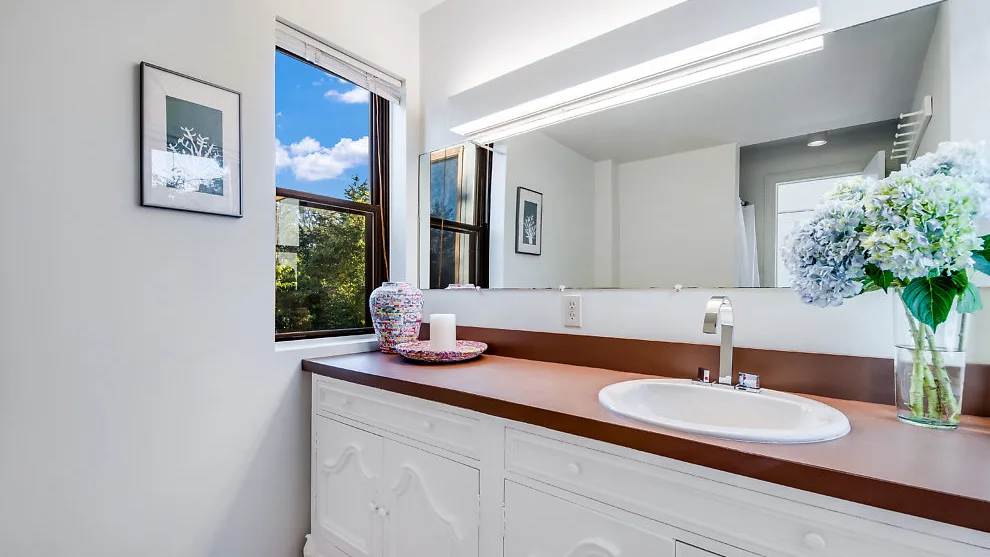 Un tocador de baño luminoso con un espejo amplio y un jarrón de flores.