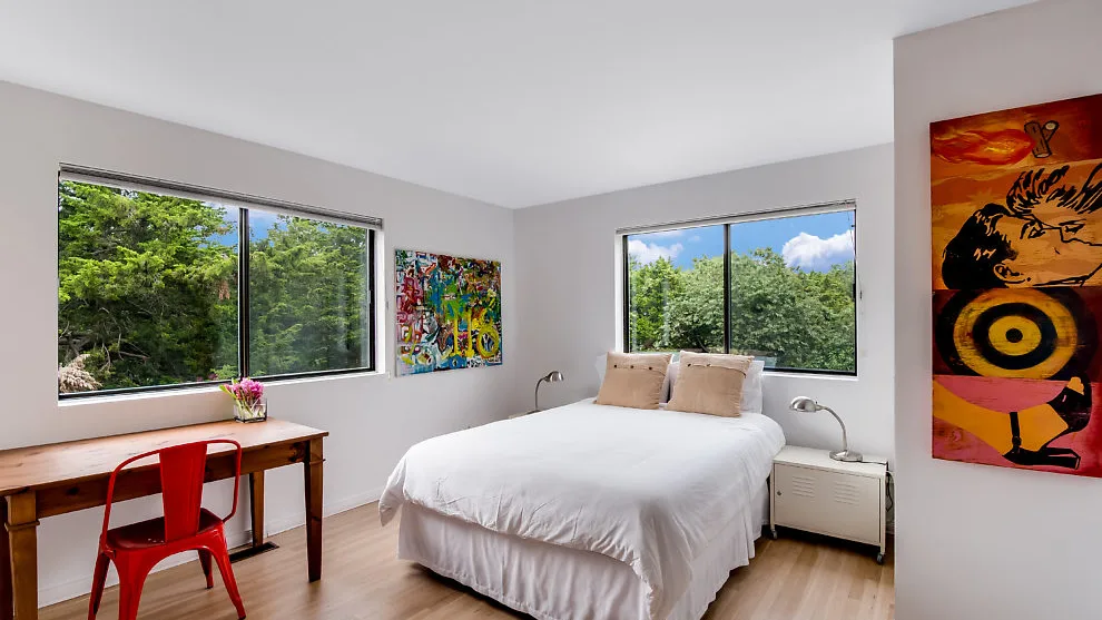 Un dormitorio luminoso con una cama, un escritorio y grandes ventanas con vistas a la vegetación.