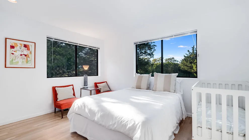 Un dormitorio luminoso con una cama, dos sillas y grandes ventanas.