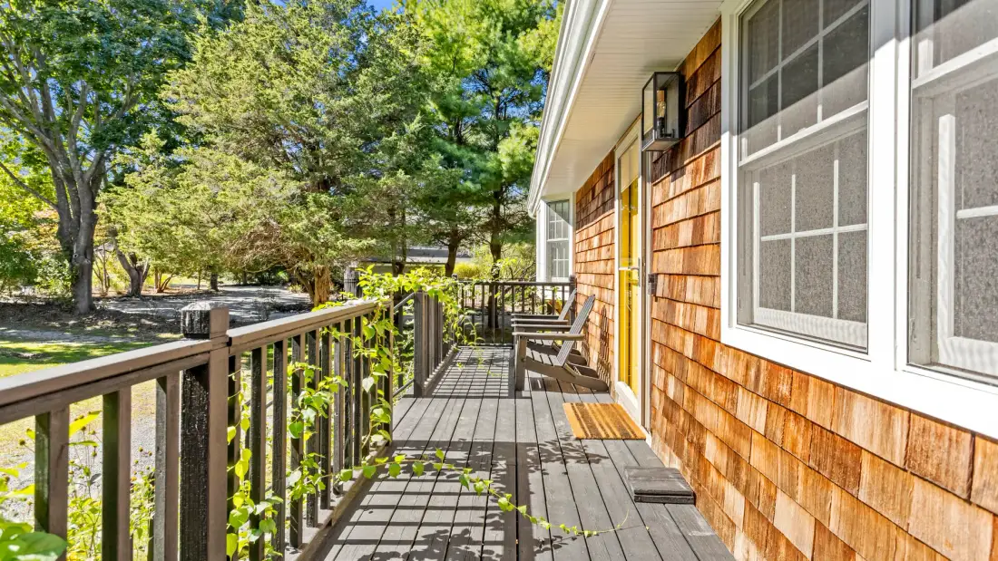 A wooden deck runs alongside a shingled house wall beneath leafy trees.