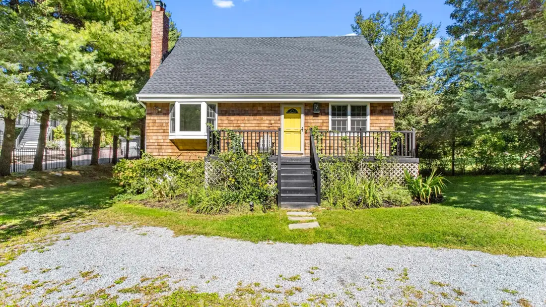 A small house with a yellow front door and a raised porch faces a gravel driveway.