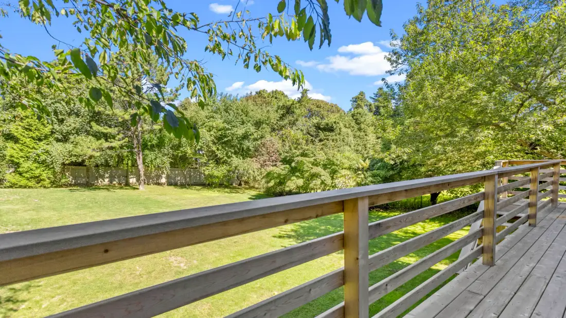 A wooden deck railing overlooks a grassy yard bordered by leafy trees.
