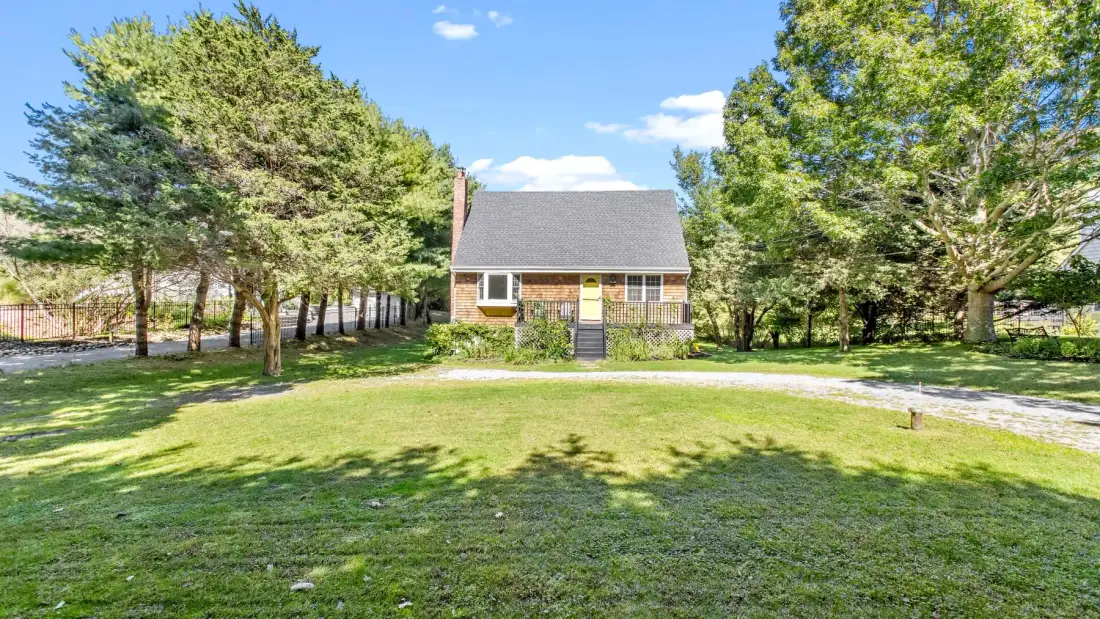 A house sits beyond a broad grassy lawn, surrounded by mature trees.