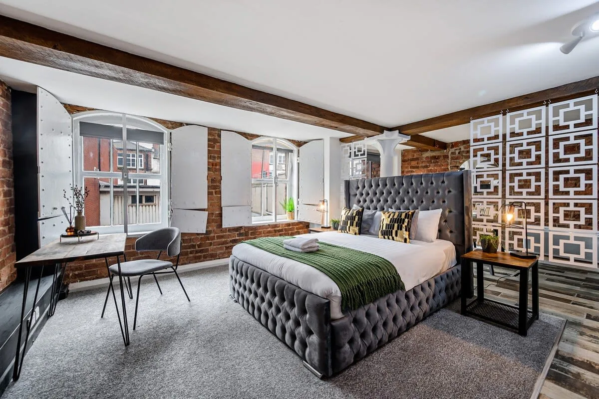 Un dormitorio con una cama tapizada grande, un escritorio y ventanas con vista a edificios cercanos.