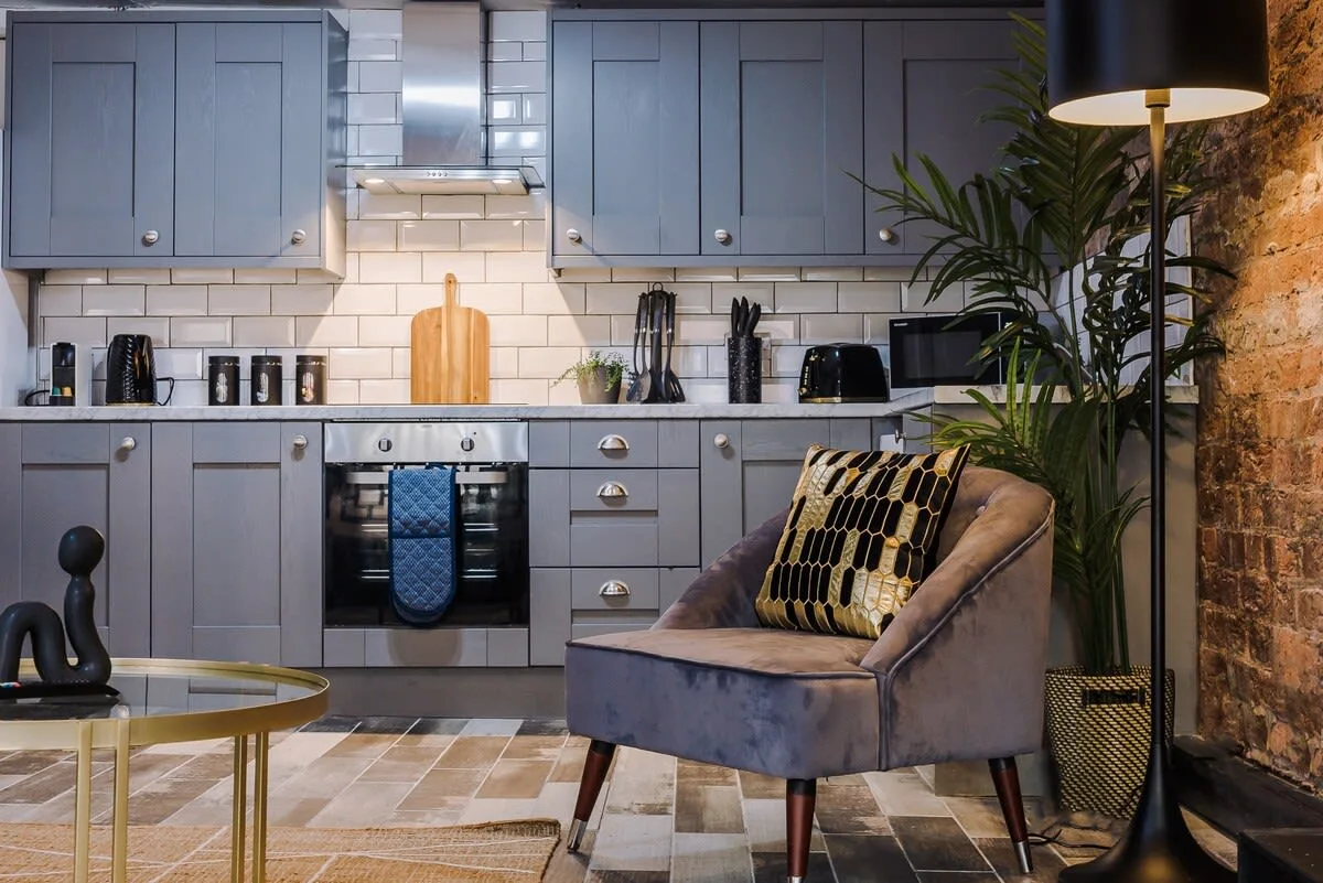 Una cocina se abre a una zona de estar con un sillón gris y una planta en maceta.