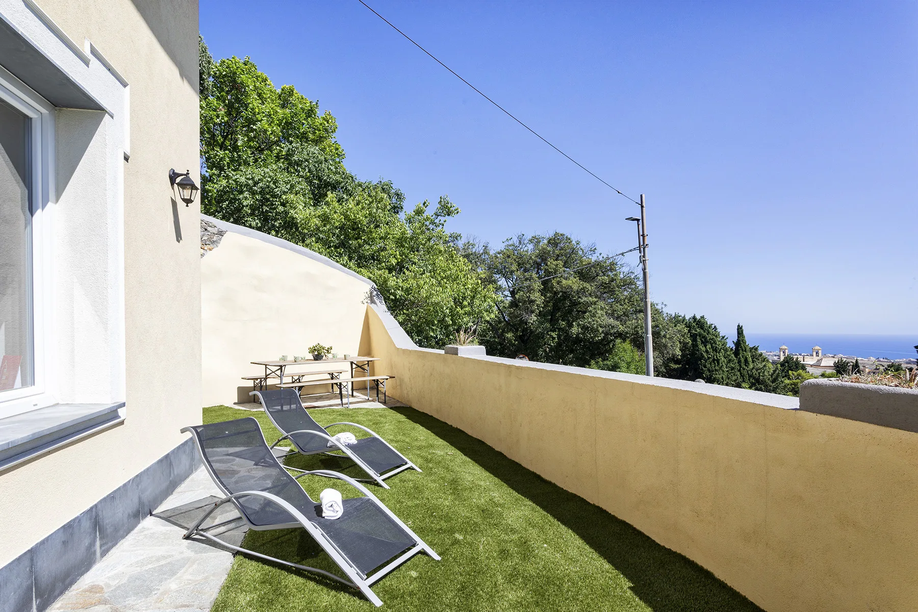 Una terraza exterior con césped tiene asientos y una mesa, con una vista costera lejana.