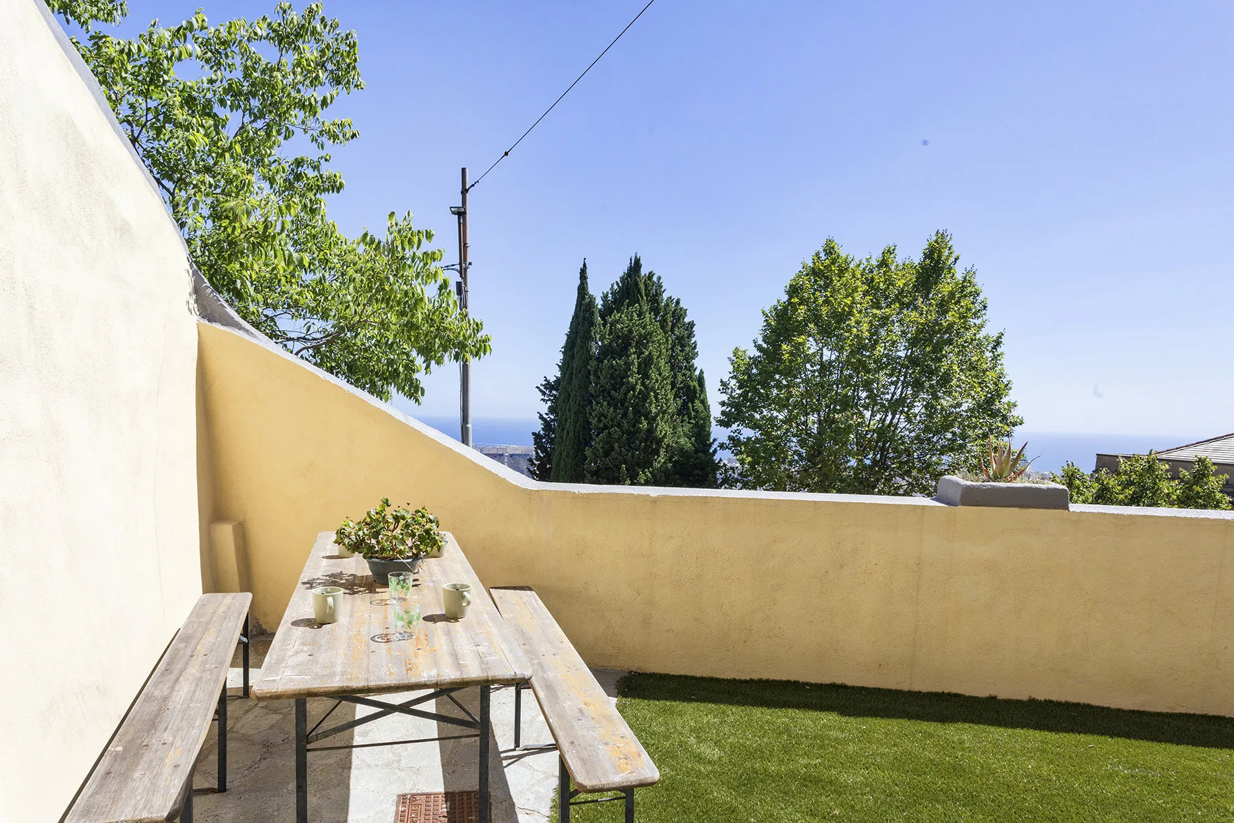 Un patio soleado al aire libre con una mesa larga, bancos y una zona de césped.