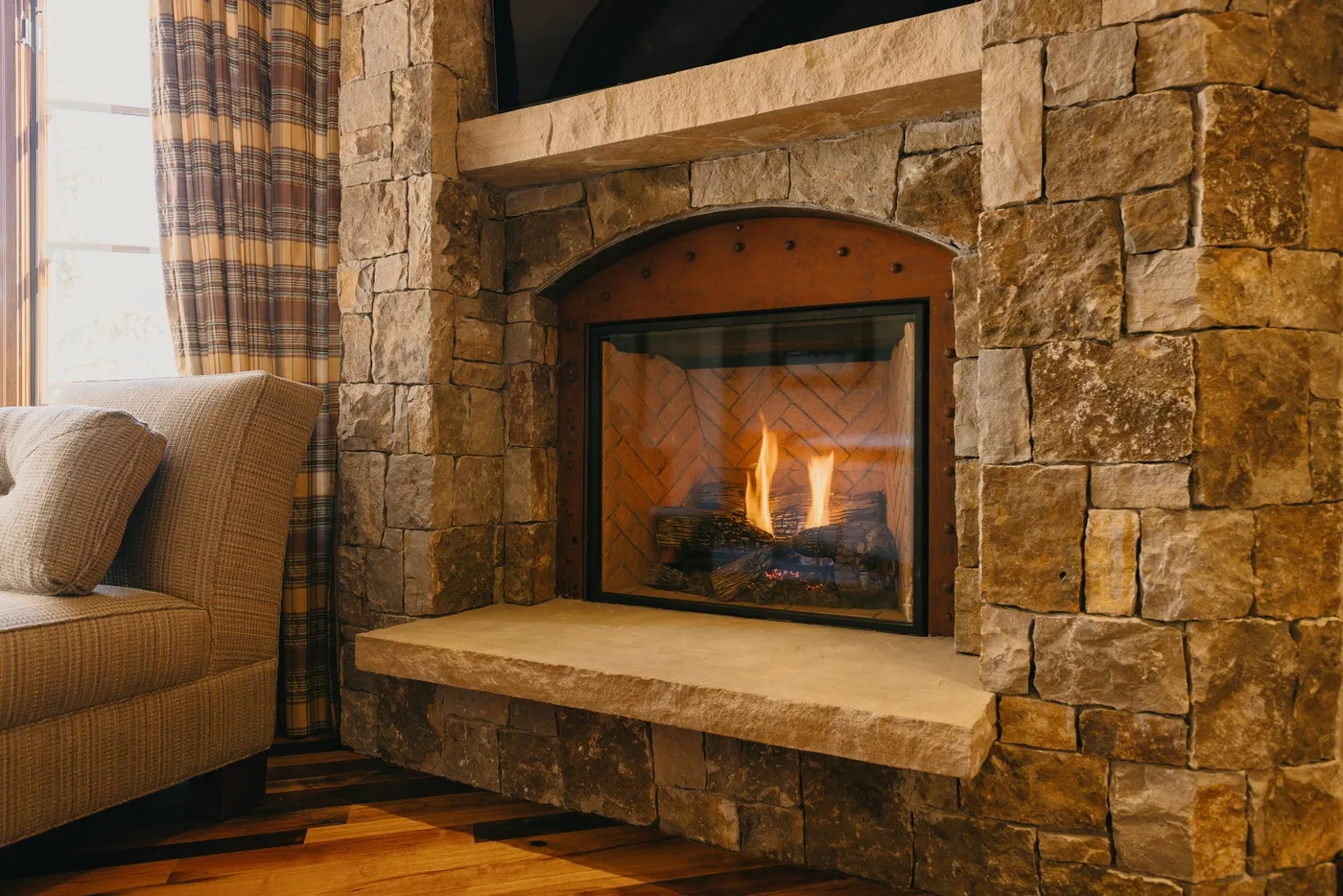 A stone fireplace with a burning fire beside a seating area and curtained window.