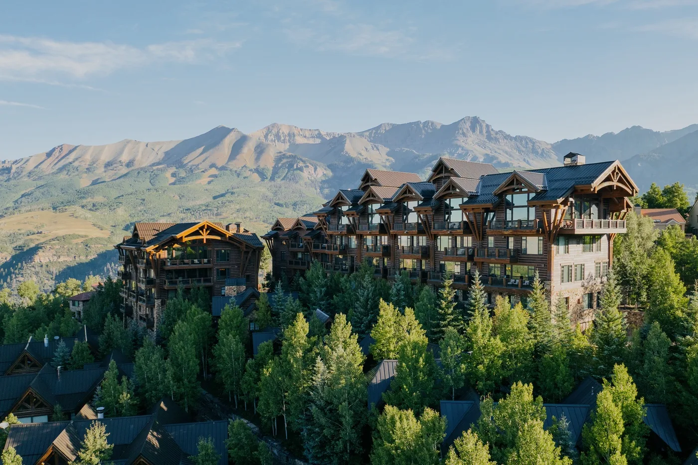 A large lodge-like building stands among trees beneath a mountain range.