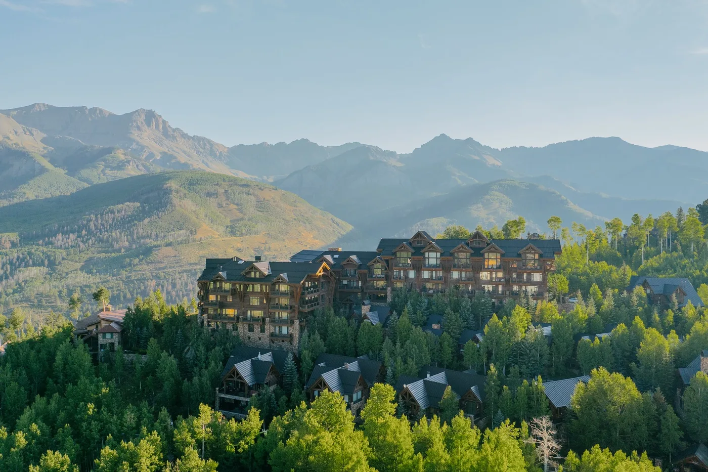 A large lodge sits among forested hills, with mountains in the background.