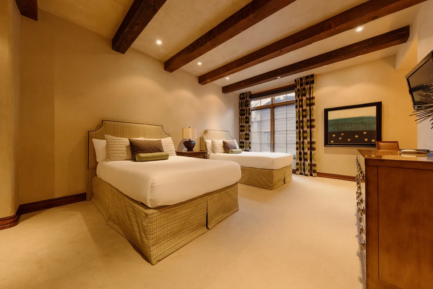 A warmly lit bedroom with two beds, exposed ceiling beams, and a window.