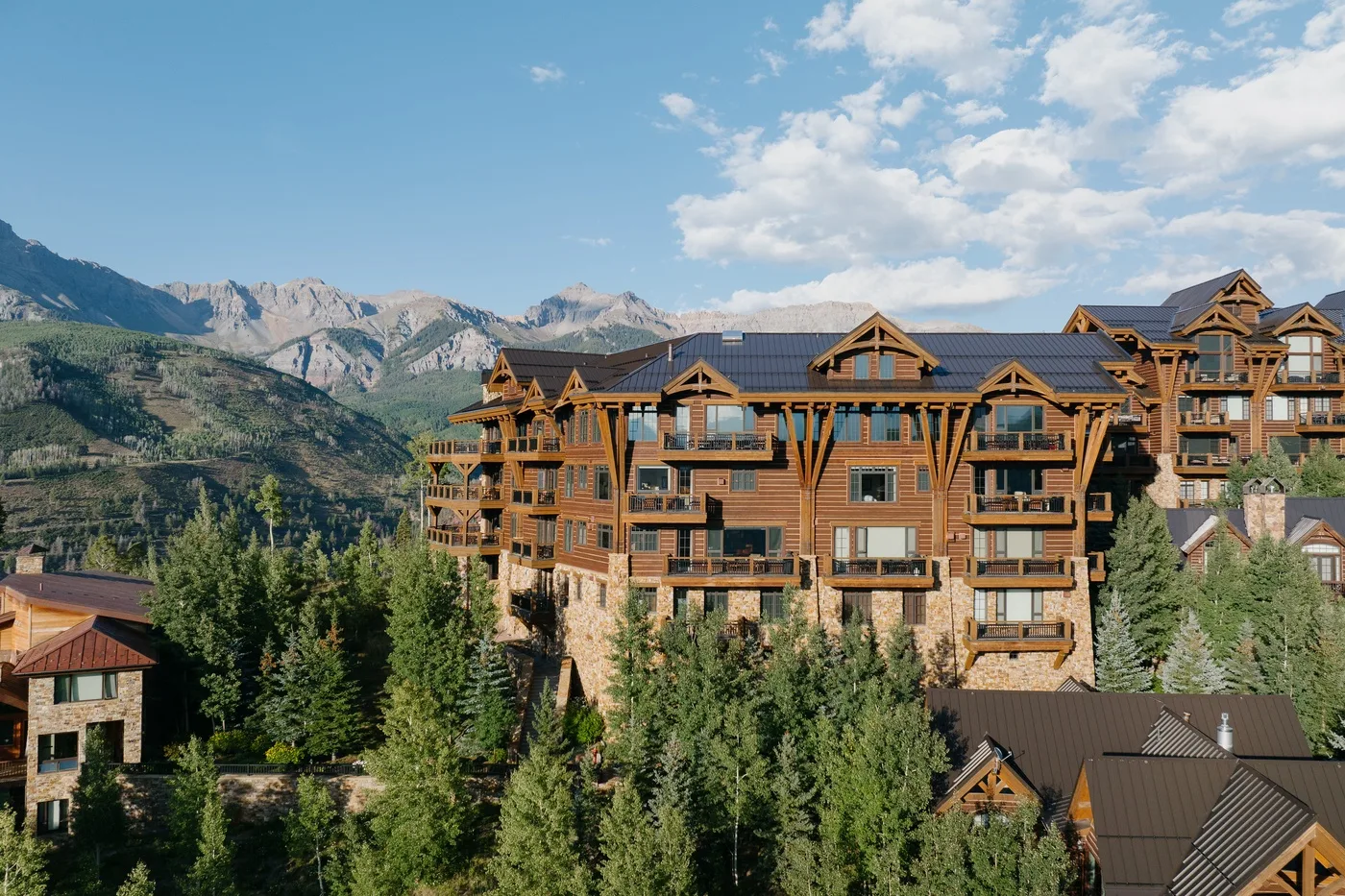 A large timber lodge stands among evergreen trees with mountains in the background.