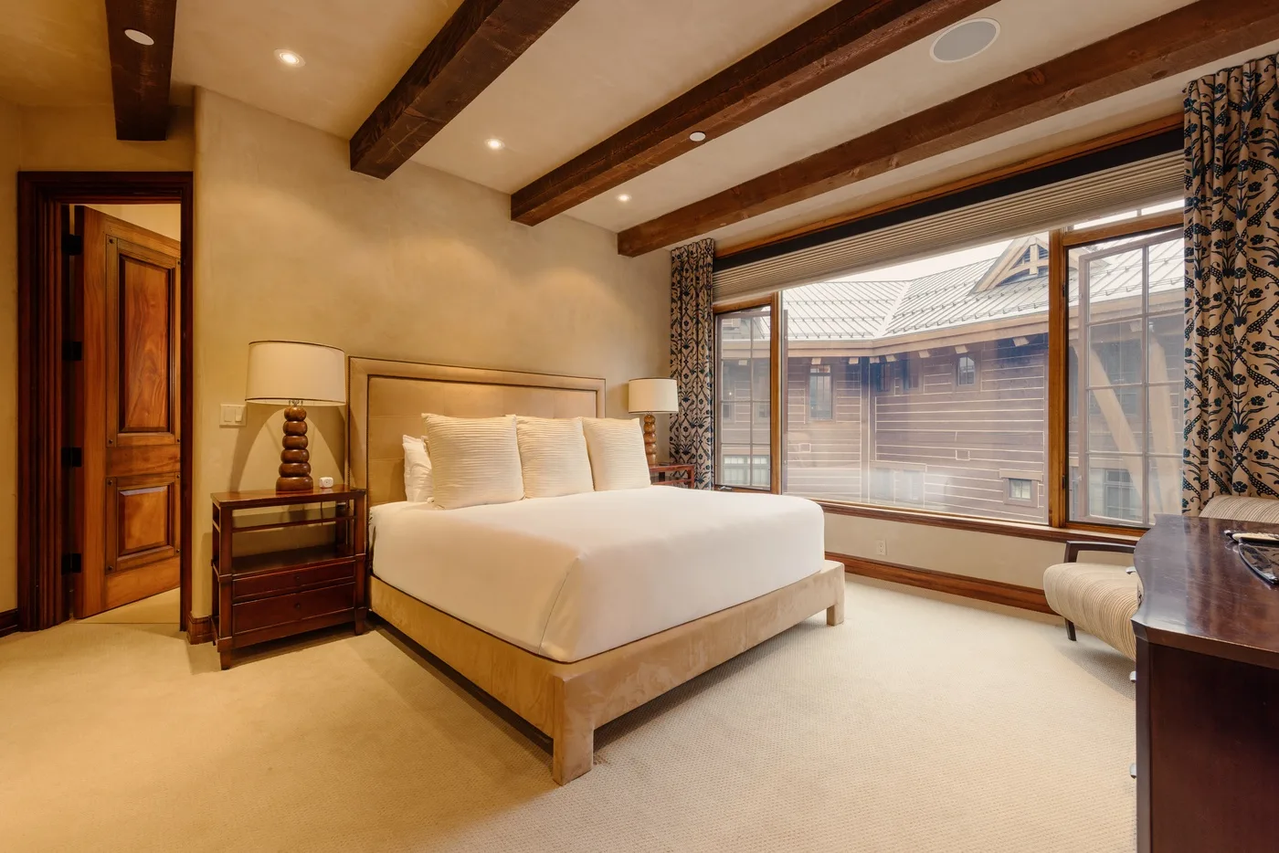 A bedroom with a bed beside large windows overlooking neighboring buildings.