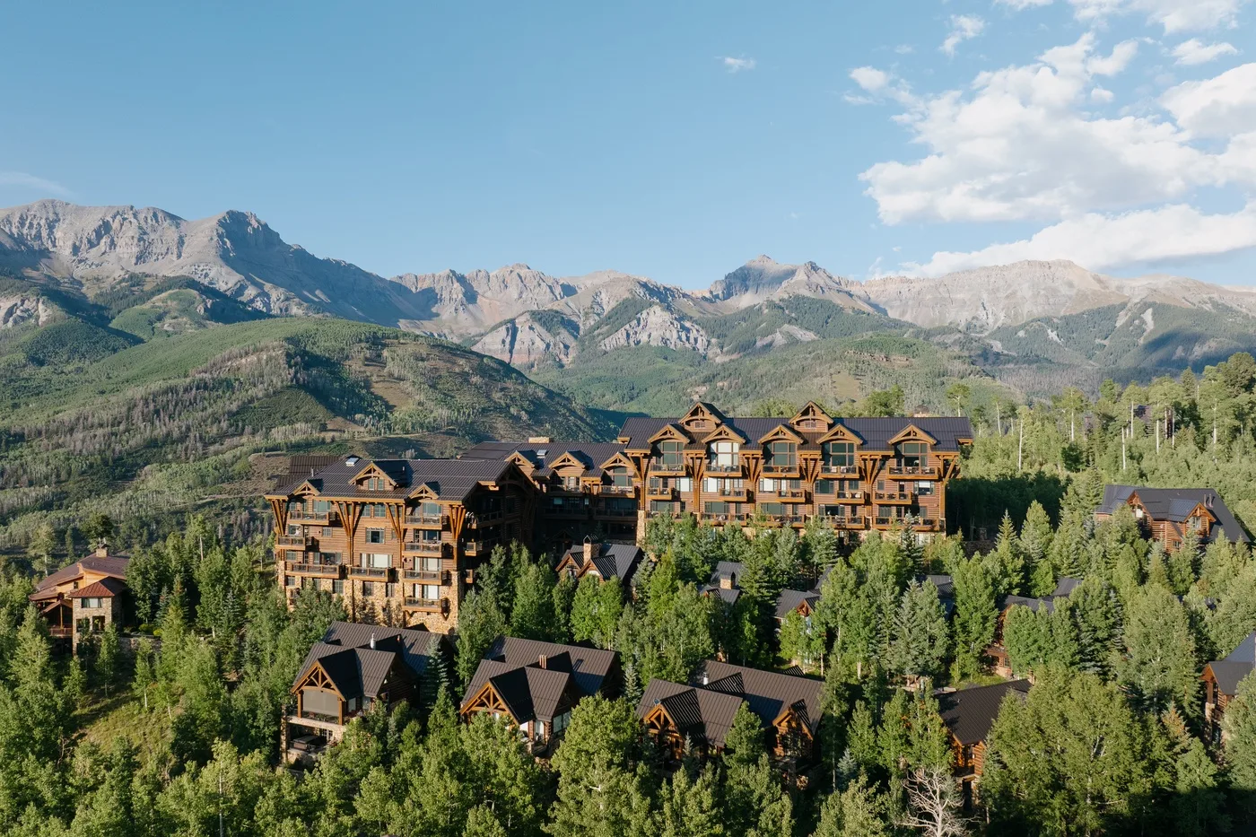 Wooden lodge buildings sit among trees beneath a mountain range and blue sky.
