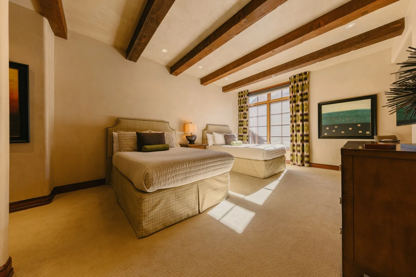 A bedroom with two beds, exposed ceiling beams, and a sunlit window.