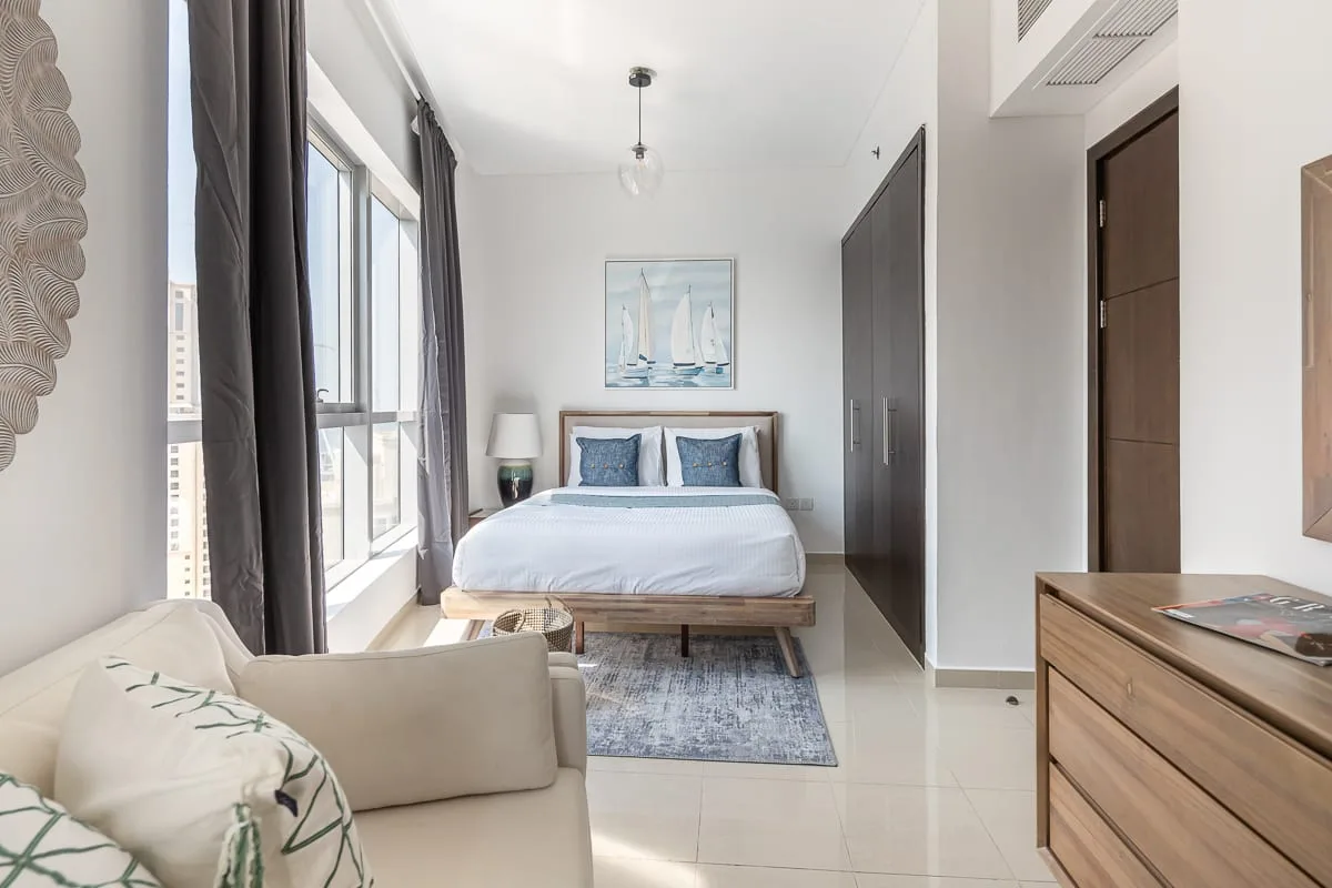 Un dormitorio luminoso con una cama junto a ventanas altas y una pequeña zona de estar.