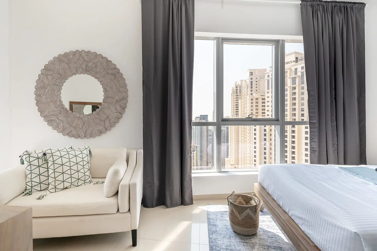 Un dormitorio luminoso con una cama, un sillón blanco y una ventana grande.