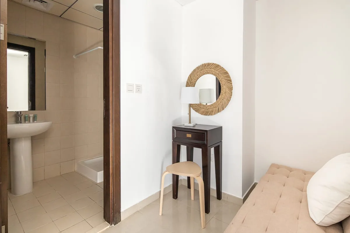 Una zona de estar del dormitorio con una mesa pequeña y un espejo redondo junto al baño abierto.