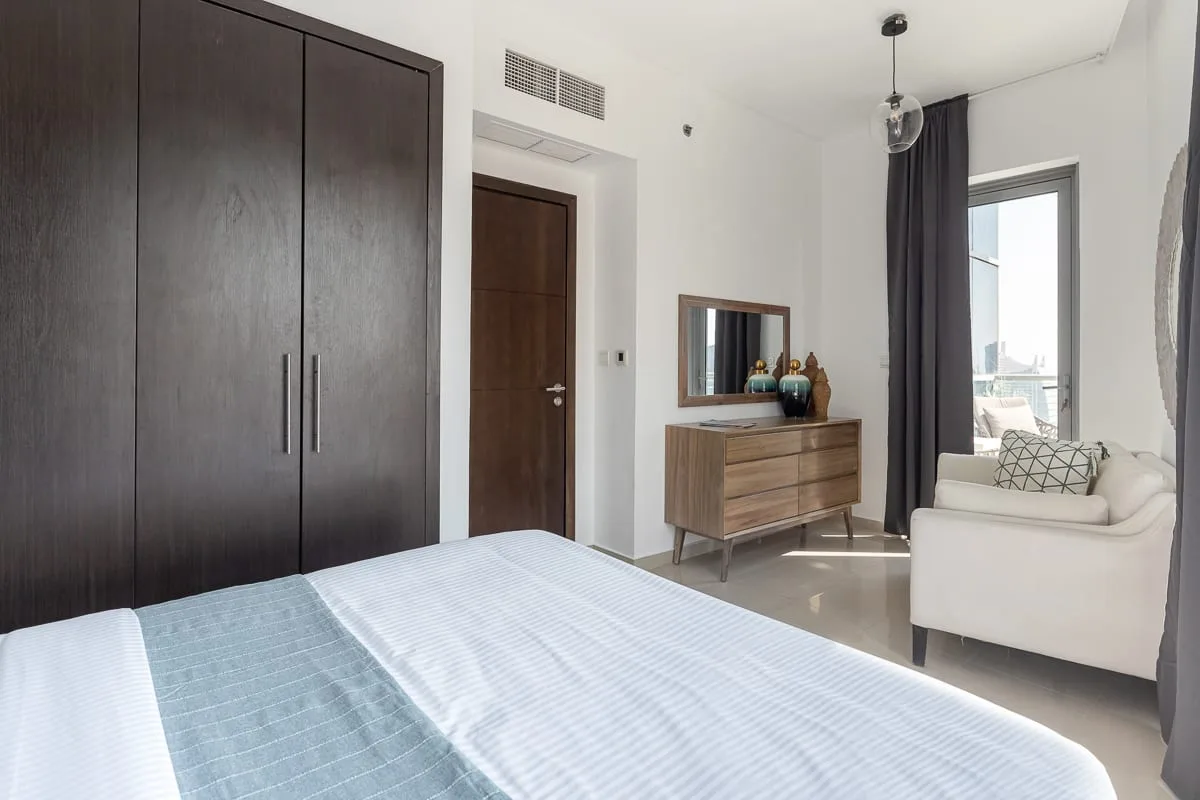 Un dormitorio luminoso con cama, armario y asientos junto a una ventana grande.