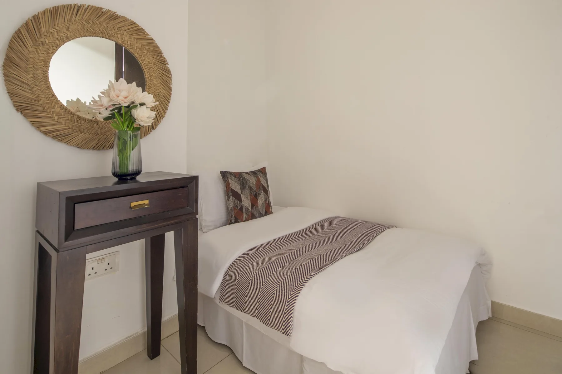 Una cama individual con ropa de cama blanca está junto a una mesa de madera oscura.