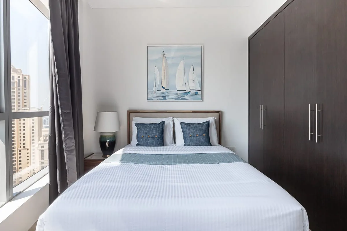 Dormitorio con una cama bien hecha, un cuadro de veleros y un armario oscuro.