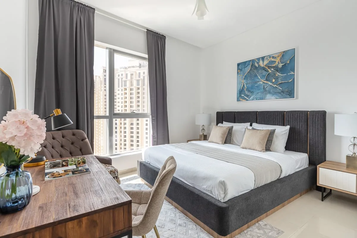Un dormitorio luminoso con una cama grande, cortinas oscuras y una ventana con vista a edificios cercanos.